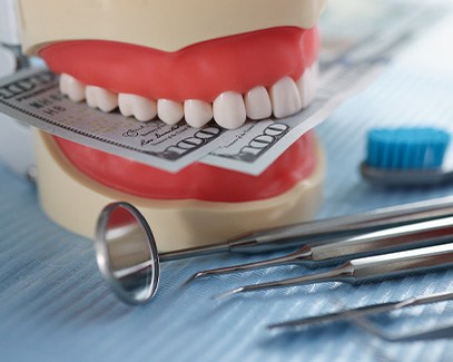 A mock jaw biting a few hundred-dollar bills next to some dental tools